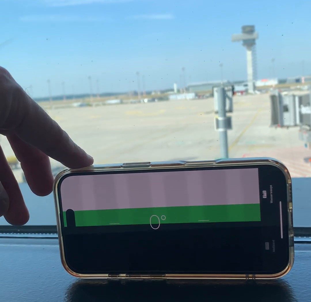 Handy mit Wasserwaage auf der Fensterbank am Flughafen BER, im Hintergrund Vorfeld und Kontrollturm, die Anzeige steht auf 0 Grad