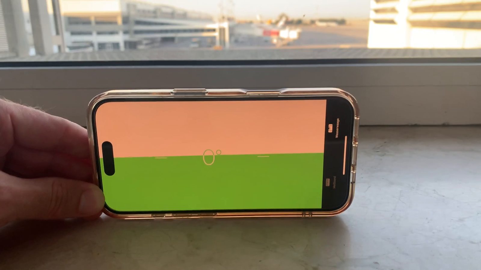 Handy mit Wasserwaage auf der Fensterbank im Terminal von Antalya, seitlich von einer Hand gehalten, draußen das Vorfeld im Abendlicht, die Anzeige steht auf 0 Grad