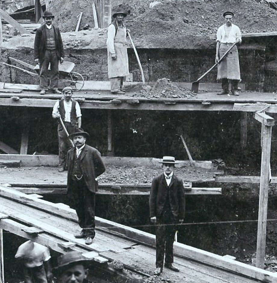 Vergrößerter Ausschnitt: Arbeiter und Herren in Anzug und Hut stehen still auf den Bohlenstegen der Rathausbaugrube 1905.